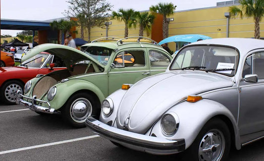 Classic silver Volkswagen Beetle on display at a Cars and Coffee event, surrounded by other vintage and modern cars. The iconic car features its signature round headlights, sleek curves, and chrome accents.