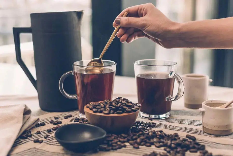 Woman stirring coffee in clear glass coffee mug on table with another filled coffee mug, cream and sugar containers, spilled roasted coffee beans, and a black pitcher.