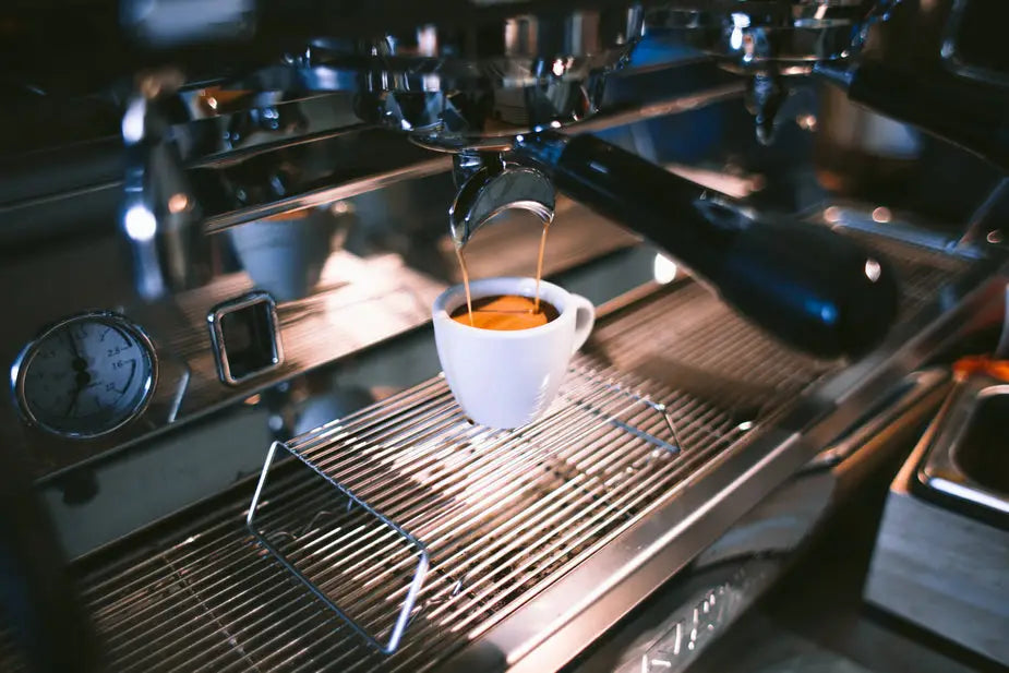 An espresso blend coffee cup sitting on a wooden surface next to a silver and black espresso machine with a steam wand and portafilter attached. The cup is filled with rich, dark espresso and the steam wand is releasing a puff of steam.